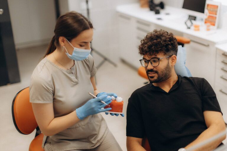 Female dentist wearing mask and gloves showing denture model to male patient explaining treatment in dental clinic